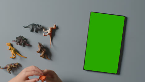 Overhead-Shot-Of-Child-Playing-With-Toy-Dinosaurs-Next-To-Green-Screen-Digital-Tablet